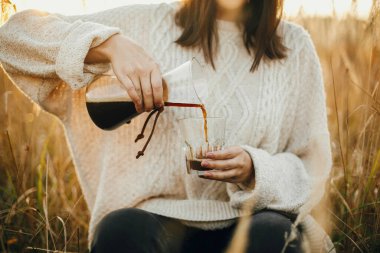 Hippi kadın günbatımında kırsal otların arka planına cam bardağa sıcak kahve dolduruyor. Alternatif kahve açık havada güneşli bir ışıkta demleniyor. Atmosferik kırsal an. Seyahat tutkusu