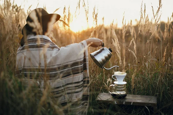 旅行中の代替コーヒー醸造 帽子のヒップスター女性は 農村部のハーブで日当たりの良い暖かい光の背景にガラスフラスコ上に注ぐフィルターでコーヒーにお湯を注ぐ — ストック写真