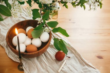 Kırsal Paskalya natürmortu. Tahta kasedeki doğal yumurtalar ve kiraz çiçeklerinin arka planındaki keten kumaş üzerindeki klasik mumlar ve kırsal masadaki yeşil yapraklar. Mutlu Paskalyalar. Organik yumurtalar
