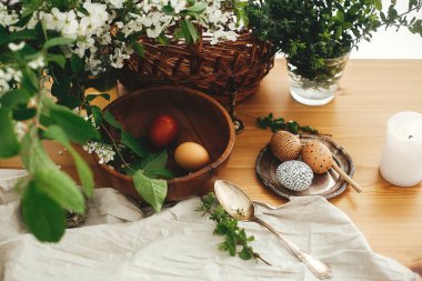 Modern balmumu süslemeleri olan şık Paskalya yumurtaları ve beyaz bahar çiçekleri, sepet, mum, keten kumaşlı sade ahşap masa üzerindeki doğal boyalı yumurtalar. Mutlu Paskalyalar. Şık kırsal yaşam. Sıfır atık