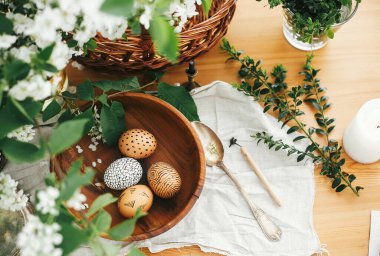 Mutlu Paskalya düzüşmeleri, sıfır israf. Hasır sepetin arka planında balmumuyla boyanmış doğal Paskalya yumurtaları beyaz bahar çiçekleri ve ahşap masadaki yeşillik. Kırsal yaşam. Eko tatil