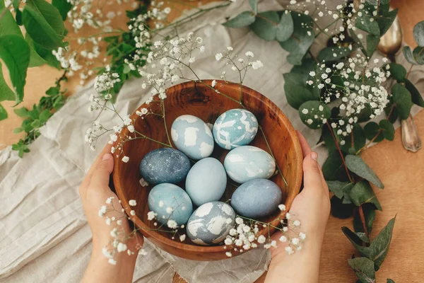 Bahar çiçeklerinde modern Paskalya yumurtaları ve okaliptüslü okaliptüsleri olan ahşap kaseyi tutan eller. Kırmızı lahanadan doğal boyayla boyanmış mavi paskalya yumurtaları. Mutlu Paskalyalar.