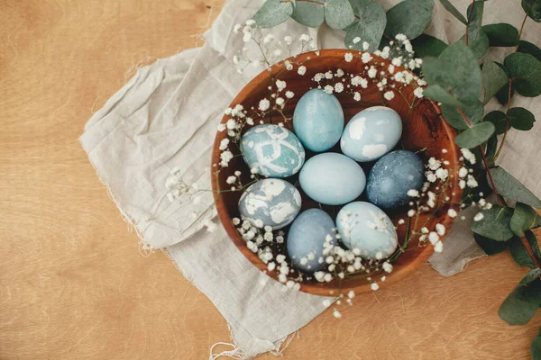 Happy Easter flat lay. Modern Easter eggs in wooden bowl with spring flowers and eucalyptus on rustic table. Stylish pastel blue Easter eggs painted in natural dye from red cabbage.