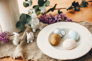 Klasik tabakta paskalya yumurtaları, beyaz tavşanlar ve ahşap masa üzerinde leylak çiçekleri. Doğal boyanmış yumurta ve bahar mor çiçeklerinin kırsal doğu kompozisyonu