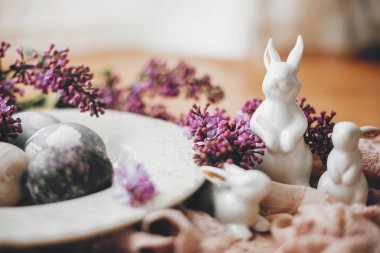 Beyaz Paskalya tavşanları, klasik tabakta modern paskalya yumurtaları ve kırsal kumaşta leylak çiçekleri. Doğal boyanmış paskalya yumurtaları ve bahar çiçekleri. Mesaj için yer var. Tatil dekorasyonu