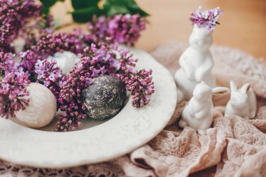 Beyaz Paskalya tavşanları, klasik tabakta modern paskalya yumurtaları ve kırsal kumaşta leylak çiçekleri. Doğal boyanmış paskalya yumurtaları ve bahar çiçekleri. Mesaj için yer var. Tatil dekorasyonu