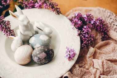 Klasik tabakta Paskalya yumurtaları, beyaz tavşanlar ve ahşap masada kumaş üzerinde leylak çiçekleri. Renkli doğal paskalya yumurtaları ve bahar çiçeklerinin kırsal bileşimi. Tatil dekorasyonu