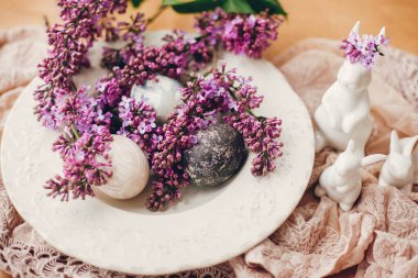 Mutlu Paskalyalar. Klasik tabakta Paskalya yumurtaları, beyaz tavşanlar ve ahşap masada kumaş üzerinde leylak çiçekleri. Renkli doğal paskalya yumurtaları ve bahar çiçeklerinin kırsal bileşimi