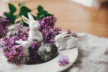 Mutlu Paskalyalar. Beyaz Paskalya tavşanları, modern paskalya yumurtası ve kırsal kumaşta leylak çiçekleri. Doğal boyanmış paskalya yumurtası ve bahar çiçekleri. Metin için boşluk.