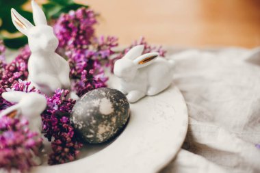 Beyaz Paskalya tavşanları, modern paskalya yumurtası ve kırsal kumaşta leylak çiçekleri. Doğal boyanmış paskalya yumurtası ve bahar çiçekleri. Mesaj için yer var. Tatil dekorasyonu