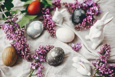 Mutlu Paskalyalar. Modern Paskalya yumurtaları, beyaz tavşanlar ve keten kumaşlı leylak çiçekleri. Şık bir tatil masası dekoru. Mesaj için yer var. Doğal boyanmış Paskalya yumurtaları ve bahar çiçekleri.