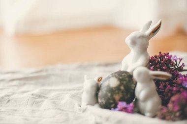 Mutlu Paskalyalar. Modern beyaz tavşanlar, paskalya yumurtası ve kırsal kumaşta leylak çiçekleri. Mesaj için yer var. Kırsal yaşam. Doğal boyanmış paskalya yumurtaları ve bahar çiçekleri. Güzel bir tatil masası dekoru.