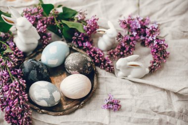 Klasik tabakta Paskalya yumurtaları, beyaz tavşanlar ve kırsal kumaş kumaşında leylak çiçekleri. Doğal boyanmış paskalya yumurtaları ve bahar çiçekleri. Tatil dekoru. Mutlu Paskalyalar