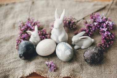 Modern Paskalya yumurtaları, beyaz tavşanlar ve kırsal kumaşta leylak çiçekleri. Şık bir tatil masası dekoru. Mesaj için yer var. Mutlu Paskalyalar. Doğal boyanmış paskalya yumurtaları ve bahar çiçekleri. Kırsal yaşam