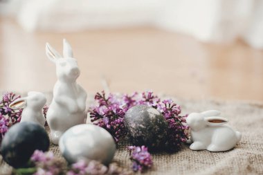 Mutlu Paskalyalar. Kırsal yaşam. Modern Paskalya yumurtaları, beyaz tavşanlar ve kırsal kumaşta leylak çiçekleri. Şık bir tatil masası dekoru. Mesaj için yer var. Doğal boyanmış Paskalya yumurtaları ve bahar çiçekleri.