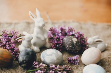 Modern Paskalya yumurtaları, beyaz tavşanlar ve kırsal kumaşta leylak çiçekleri. Şık bir tatil masası dekoru. Mesaj için yer var. Mutlu Paskalyalar. Doğal boyanmış paskalya yumurtaları ve bahar çiçekleri. Kırsal yaşam