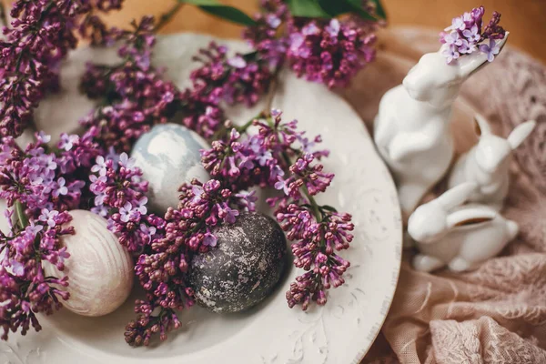 复活节快乐老式盘子上有时尚的复活节彩蛋 木制桌子上有白兔和紫丁香花 农村五彩缤纷的天然色彩艳丽的彩蛋和春花组合 — 图库照片