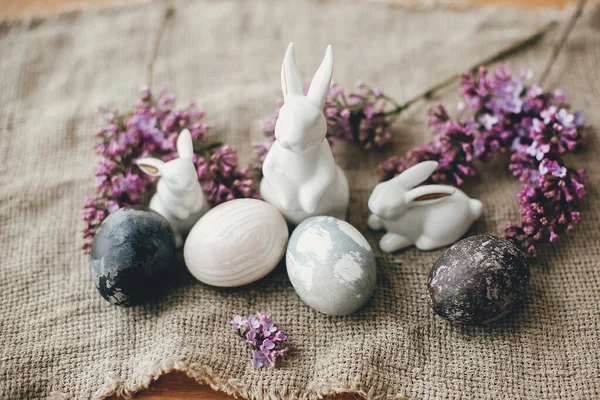 Modern Easter eggs, white bunnies and lilac flowers on rustic fabric. Stylish holiday table decor. Space for text. Happy Easter. Natural dyed easter eggs and spring flowers. Rural still life