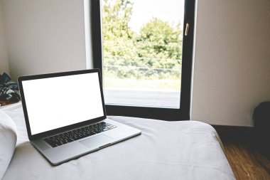 Modern odadaki büyük pencerenin arka planında boş beyaz ekranlı dizüstü bilgisayar, kopyalama alanı. Ev modelinden çevrimiçi çalışıyor. Serbest meslek kavramı. İnternetten alışveriş. Evde kalıyorum. Günlük şablonu
