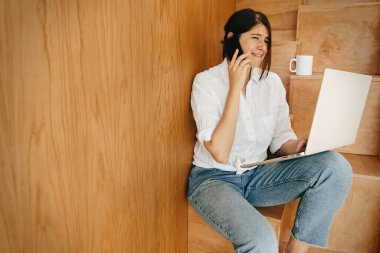 Komik duyguları olan mutlu bir kız telefonda konuşuyor, dizüstü bilgisayar kullanıyor modern odada ahşap merdivenlerde oturuyor. İnternetten alışveriş yapan ya da internetten çalışan genç bir kadın. Evde kal.