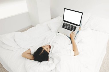 Beyaz modern odadaki boş ekranlı, dizüstü bilgisayarla yatakta yatan mutlu stil kızı. Genç bir kadın boş ekrana bakıyor, online alışveriş yapıyor ya da ev şablonundan online çalışıyor. Bağımsız