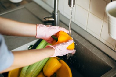 Sebze yıkıyorum. Eldivenli eller virüs salgını sırasında lavaboda su altında biber yıkıyor. Pembe ellerdeki kadın taze sebzeleri temizliyor, modern mutfakta yemek yapmaya hazırlanıyor.