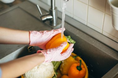 Eldivenli eller virüs salgını sırasında lavaboda su altında biber yıkıyor. Sebze yıkıyorum. Pembe ellerdeki kadın taze sebzeleri temizliyor, modern mutfakta yemek yapmaya hazırlanıyor.
