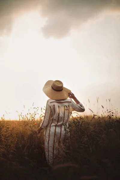 素朴なドレスと帽子の女性は 夏の牧草地で野の花やハーブで平和な夕日を楽しんでいます 大気中の本物の瞬間 夕方田舎を歩くスタイリッシュな女の子 農村のスローライフ — ストック写真