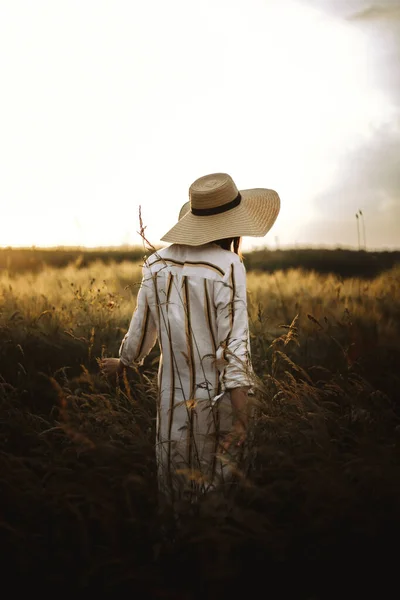 素朴なドレスと帽子の女性は 夏の牧草地で野の花やハーブで平和な夕日を楽しんでいます 大気中の本物の瞬間 夕方田舎を歩くスタイリッシュな女の子 農村のスローライフ — ストック写真