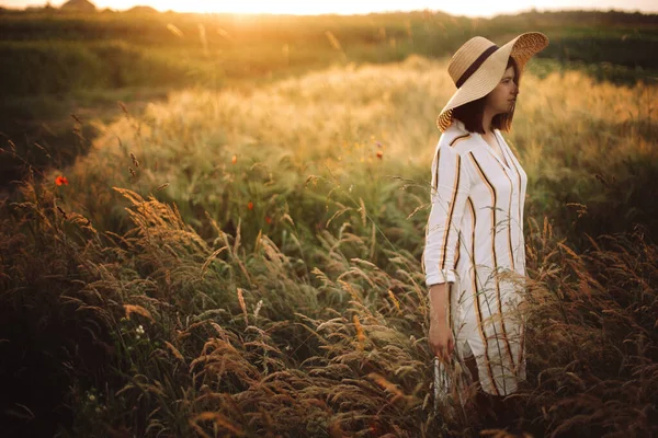 在夏日的草地上 身穿乡村服装 头戴帽子 头戴野花 头戴香草 在夕阳西下金光闪闪的暮色中散步的女人 时髦的女孩在乡村享受夜晚 农村缓慢的生活 复制空间 — 图库照片