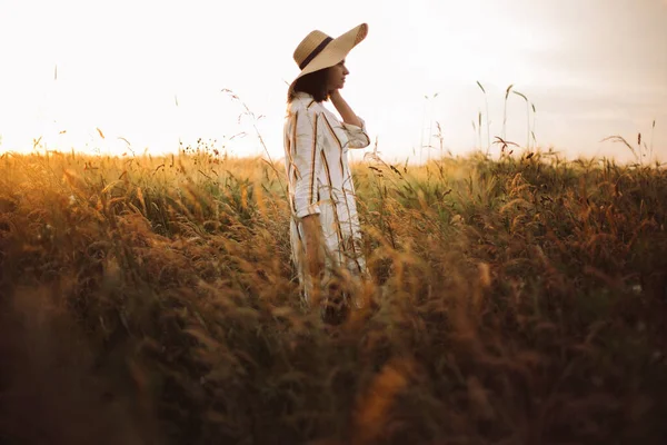 素朴なドレスと帽子の女性は 夏の牧草地で日没の黄金の光の中で野の花やハーブの中を歩く 田舎で夜を楽しむスタイリッシュな女の子 農村部のスローライフ スペースのコピー — ストック写真