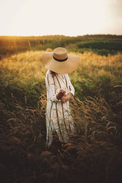素朴なドレスと帽子の女性は 夏の牧草地で日没の黄金の光の中で野の花やハーブの中を歩く 大気中の本物の瞬間 田舎で夜を楽しむスタイリッシュな女の子 — ストック写真