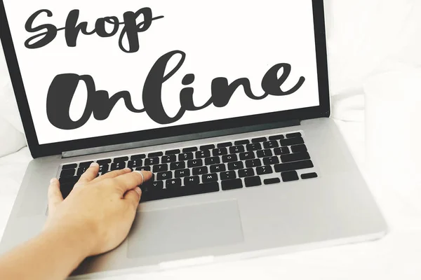 Shop online text. Shopping online lettering on laptop screen. Person using laptop on white cozy bed. Shop online from home, hand on keyboard. Isolation at home. Stay home