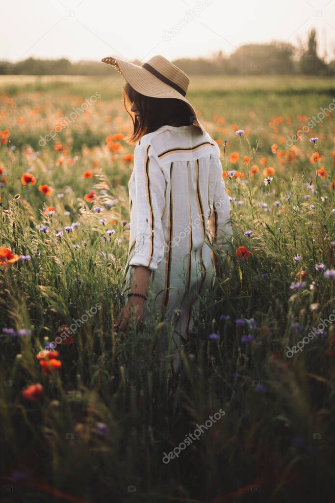 Stylish girl in hat walking in wildflowers in sunset light in summer meadow. Young woman in linen dress walking among poppy and cornflowers in countryside. Rural slow life. Enjoying simple life