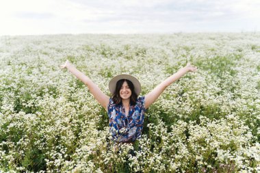 Mutlu genç kadın yaz çayırlarında kır çiçeklerinde gülümsüyor. Mavi elbiseli ve şapkalı güzel, neşeli kız tarlada eğleniyor, mutluluk anı.