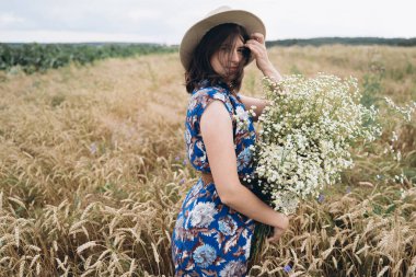 Kırsalda sakin bir yaz. Buğday tarlasında beyaz kır çiçekleriyle poz veren mavi elbiseli ve şapkalı şık genç bir kadın. Büyük papatya desenli güzel bir kızın şehvetli portresi..