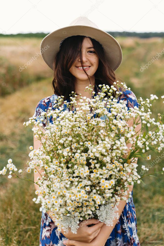 Stylish young woman in blue vintage dress and hat walking with white wildflowers in summer meadow. Happy beautiful girl smiling with daisies bouquet in field. Summer in countryside
