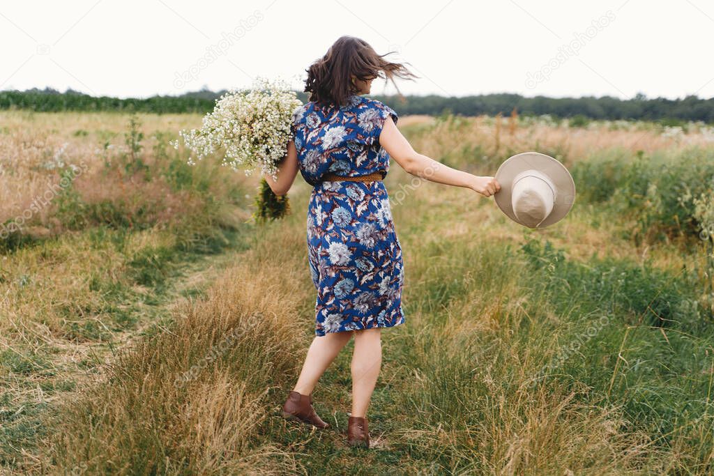 Young woman in blue vintage dress and hat dancing with white wildflowers in meadow. Beautiful girl having fun and laughing in windy field with big daisies bouquet. Authentic summer in countryside