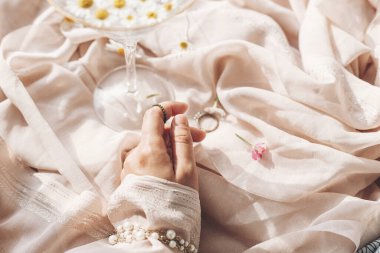 Yumuşak bej kumaşın arka planında mücevherler, şık bir şarap kadehi ve suda papatya çiçekleri. Yumuşak çiçekli estetik. Yaratıcı yaz imajı. Bohem havası.