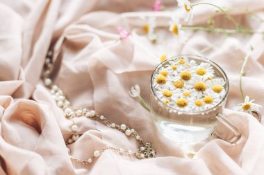 Yumuşak bej kumaş, kır çiçekleri ve mücevherlerle kaplı cam bardaktaki Daisy çiçekleri. Yumuşak çiçekli estetik. Metin için alanı olan yaratıcı bir yaz görüntüsü. Bohem havası.