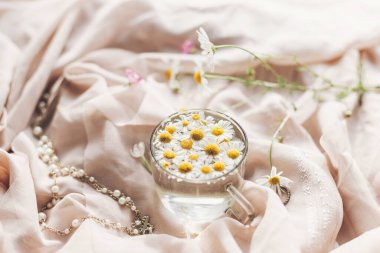 Yumuşak çiçekli estetik. Yumuşak bej kumaş, kır çiçekleri ve mücevherlerle kaplı cam bardaktaki Daisy çiçekleri. Metin için alanı olan yaratıcı bir yaz görüntüsü. Bohem havası.