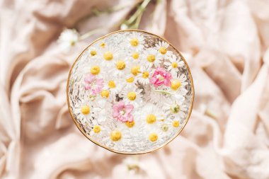 Yumuşak çiçekli estetik. Metin için alanı olan yaratıcı bir yaz görüntüsü. Bohem havası. Yumuşak bej kumaşın arka planında, şık şarap kadehinde sudaki kır çiçekleri. Üst görünüm