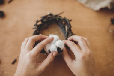 Çiçekçi, kır yılbaşı mum tutacağı yapıyor. Elleri pamuk tutuyor ve ağaç dalları ve çam kozalaklarıyla ahşap masaya kırsal şenlik dekorasyonu yapıyor. Tatil atölyesi