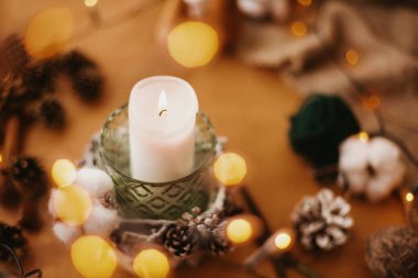 Noel mumu, ağaç dalları, pamuk, anason ve kozalaklardan yapılmış kır evinde, şenlikli ışıklar altında, ahşap masanın üzerinde. Kırsal Noel mum dekoru yapıyorum. Tatil atölyesi