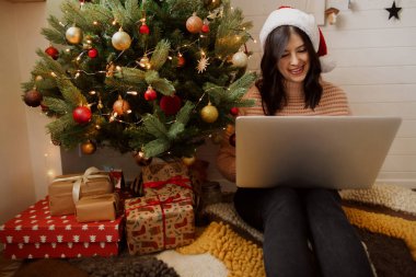 Noel Baba şapkalı mutlu kız dizüstü bilgisayarla çalışıyor, Noel alışverişi yapıyor. Modern odada dizüstü bilgisayarla yılbaşı ağacının altında ışıklar ve süslemelerle oturan zarif genç bir kadın..