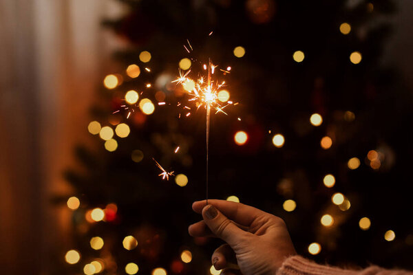 Burning sparkler in hand on background of golden bokeh lights in festive dark room. Happy New Year. Hand holding firework at christmas tree with golden illumination.
