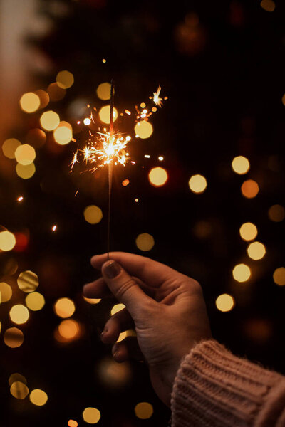 Burning sparkler in hand on background of golden bokeh lights in festive dark room. Happy New Year. Hand holding firework at christmas tree with golden illumination.