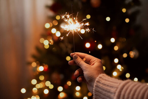 Happy New Year. Burning sparkler in hand on background of golden bokeh lights in festive dark room. Hand holding firework at christmas tree with golden illumination.