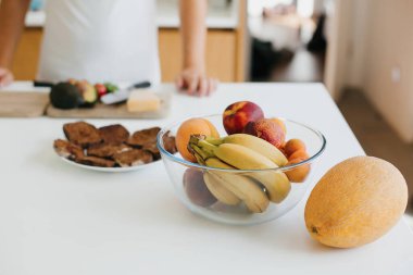 Arka planda meyveler olan şık cam kase avokado, kiraz domatesli tost, beyaz modern mutfakta ekmek yapıyor. Muz, şeftali, kayısı ve kavun tezgahın üstünde.