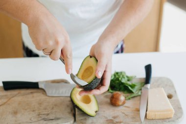 Modern beyaz mutfakta mükemmel olgunlaşmış avokadoyu kaşıkla soyan kişi. Avokado, domates, roka ve peynirle sağlıklı tost yapma süreci. Ev yemeği konsepti